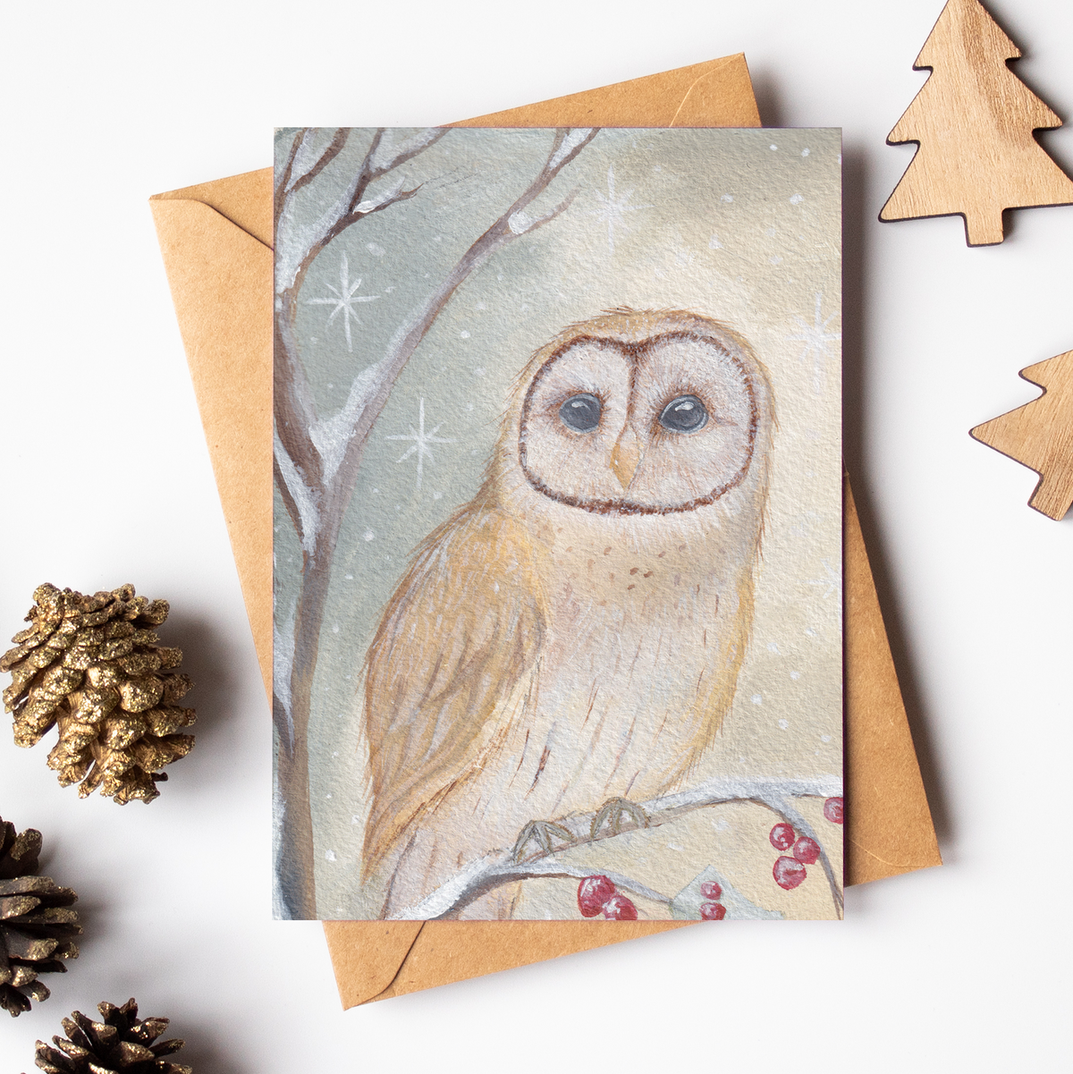 A square greeting card featuring a painted barn owl in a snowy winter scene, placed on top of a kraft paper envelope. The card is styled on a white background with wooden Christmas tree cutouts, pinecones, and a small wooden ornament.