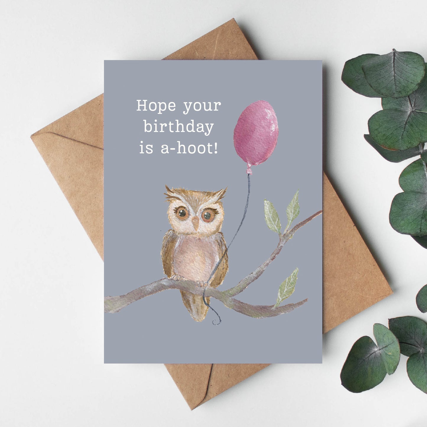 Greeting card with owl and balloon illustration, envelope, and eucalyptus leaves on a light background