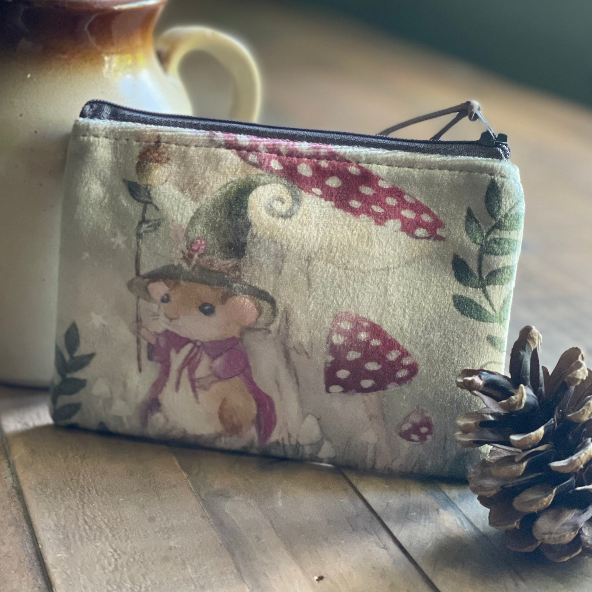 Small pouch with a bear and mushroom design on a wooden surface next to a vase and pine cone.