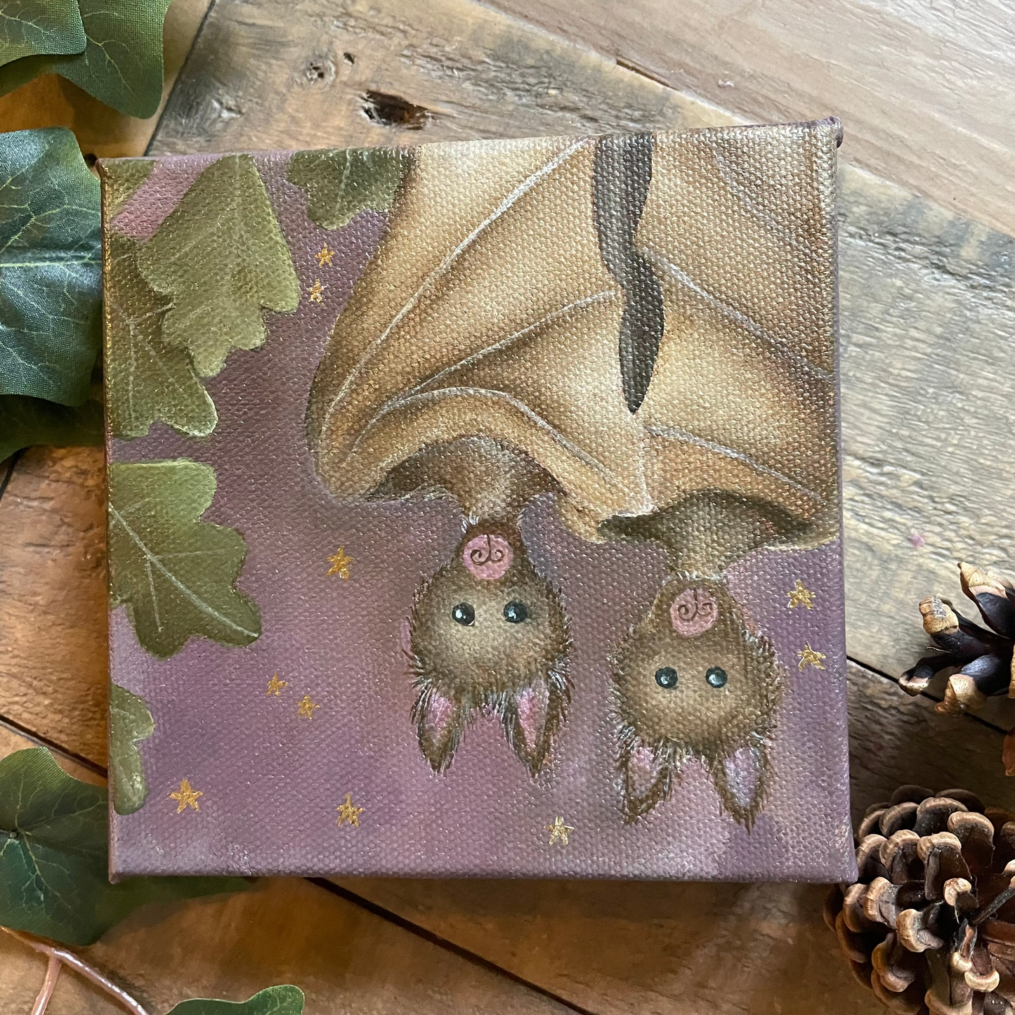 Canvas print of a bat and two baby bats on a purple background with green leaves and pinecones around.