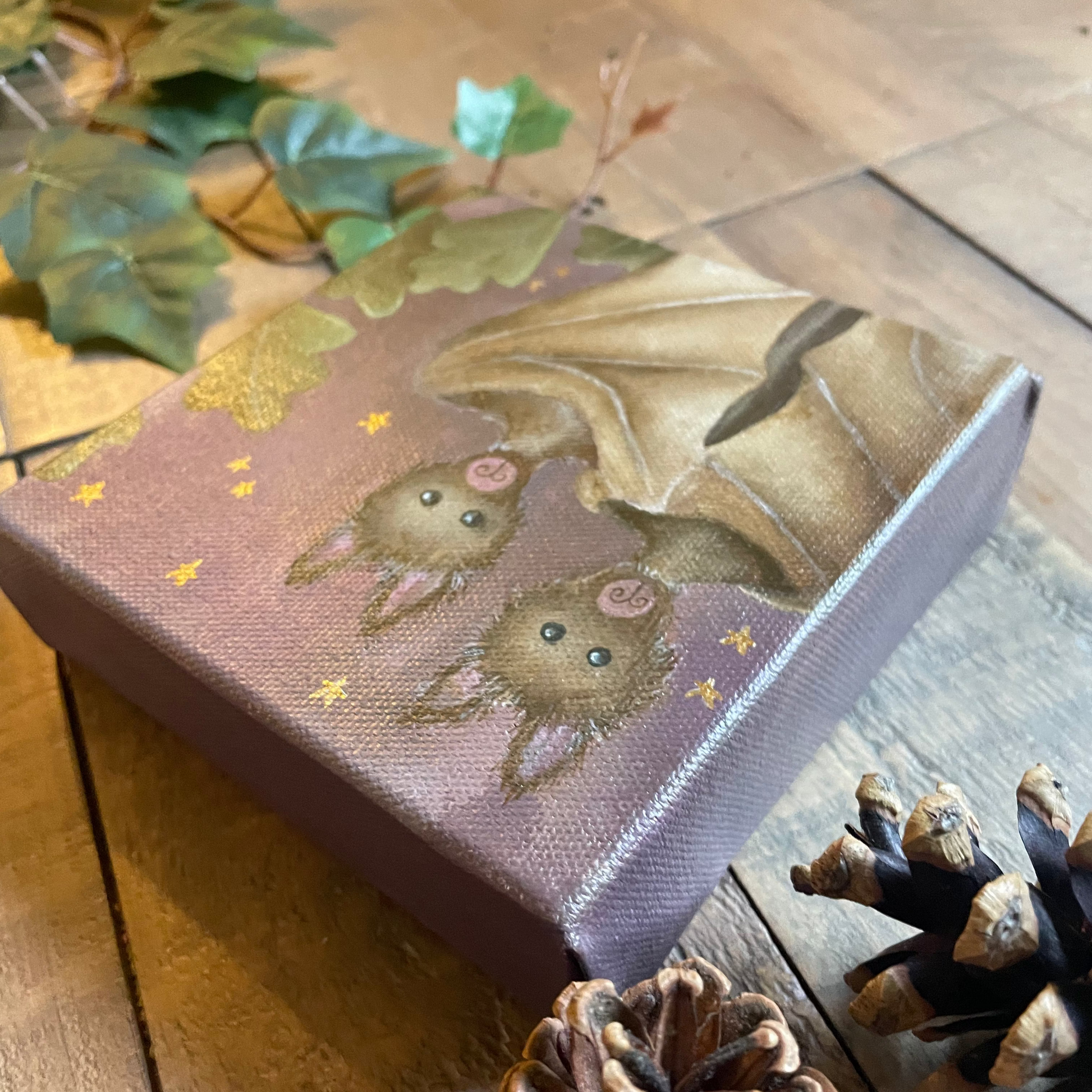 Small rectangular box with embroidered design of a bat and leaves on a wooden surface with pine cones.