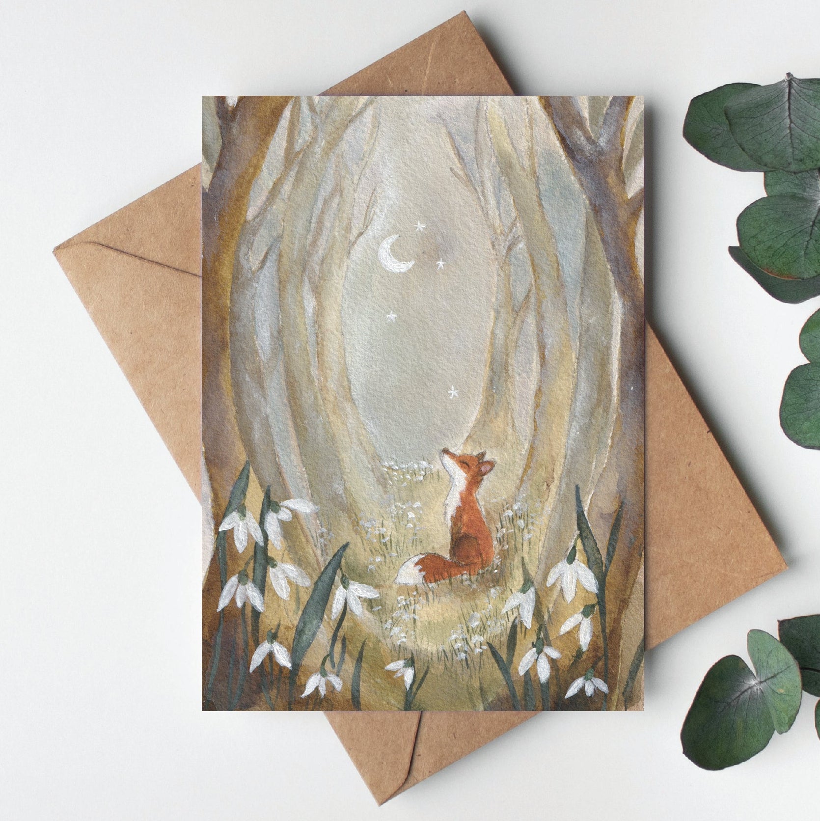 Greeting card with a fox in a forest design, placed on a white surface with eucalyptus leaves.