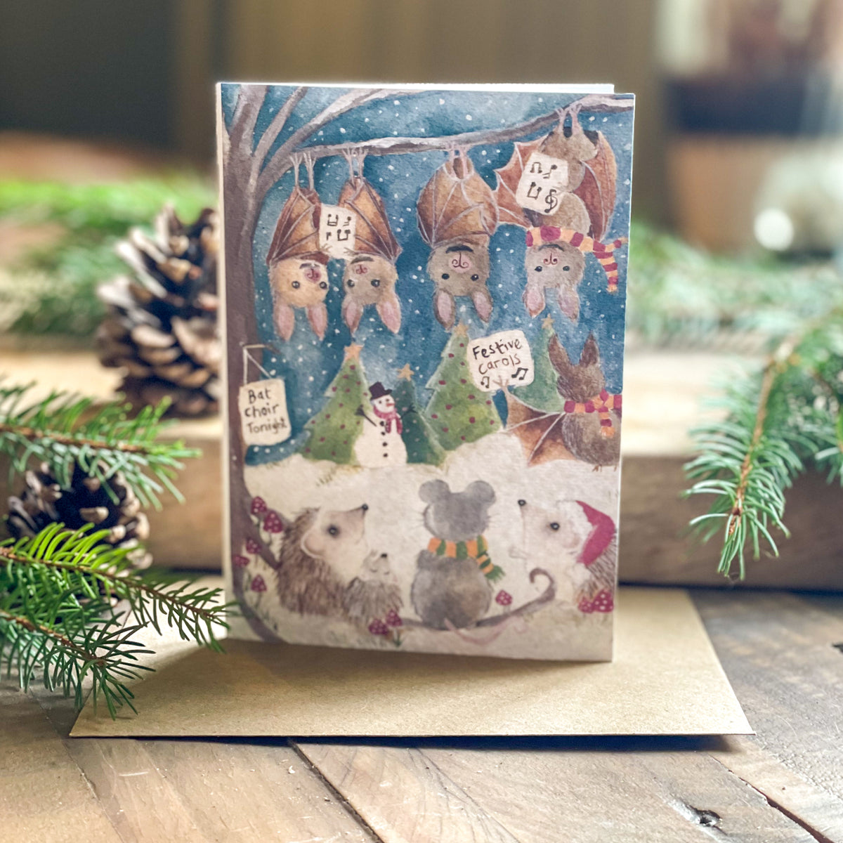 A Christmas-themed illustrated greeting card featuring bats and mice in a snowy winter scene, displayed on a kraft envelope with pine branches and a pinecone around it.