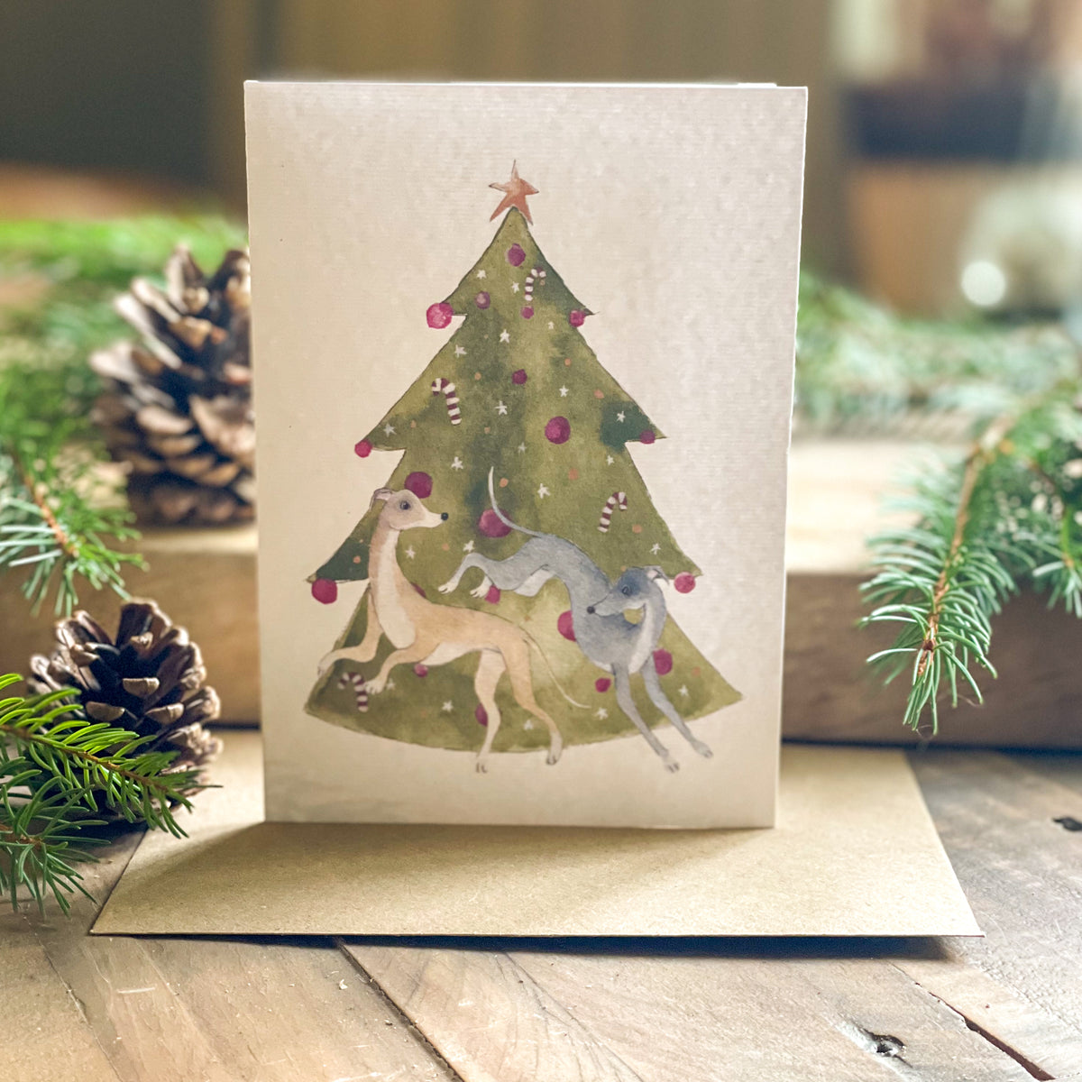 A handmade Christmas greeting card featuring a watercolor illustration of a green Christmas tree with ornaments and two dogs, displayed on a wooden surface with pine branches and a kraft envelope.