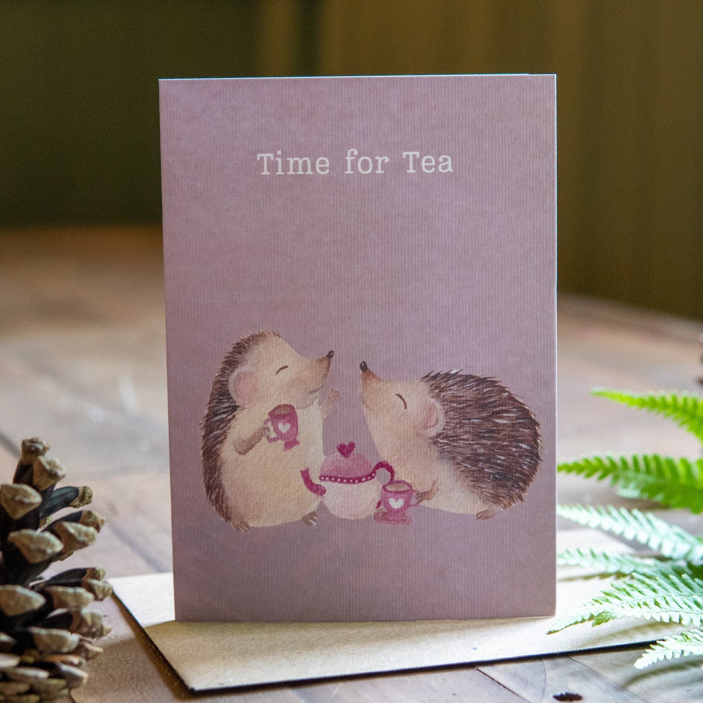 Greeting card with hedgehogs and 'Time for Tea' text on a wooden surface with plants.