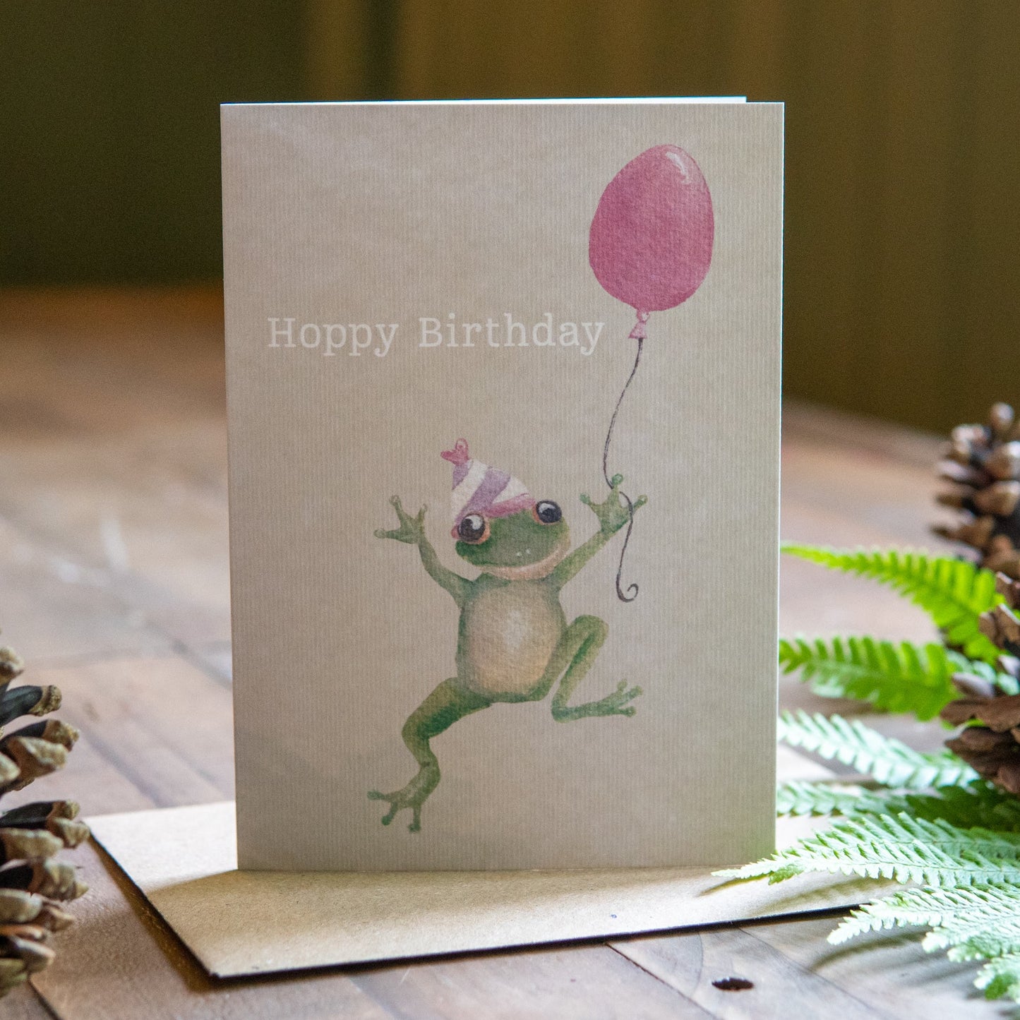 Birthday card with a frog holding a pink balloon on a wooden surface with pine cones and ferns.