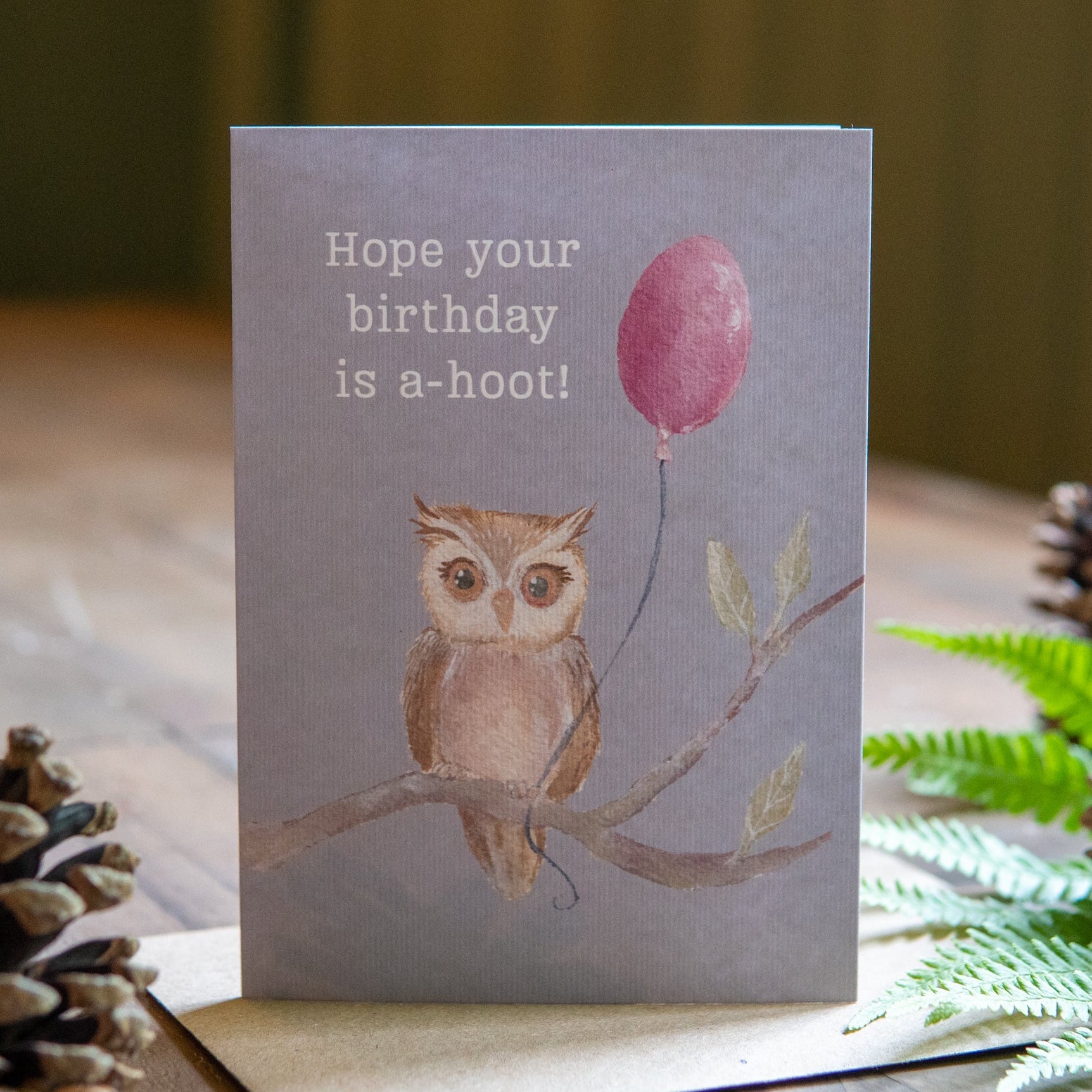 Greeting card with owl and balloon illustration on a wooden surface with decorative elements.