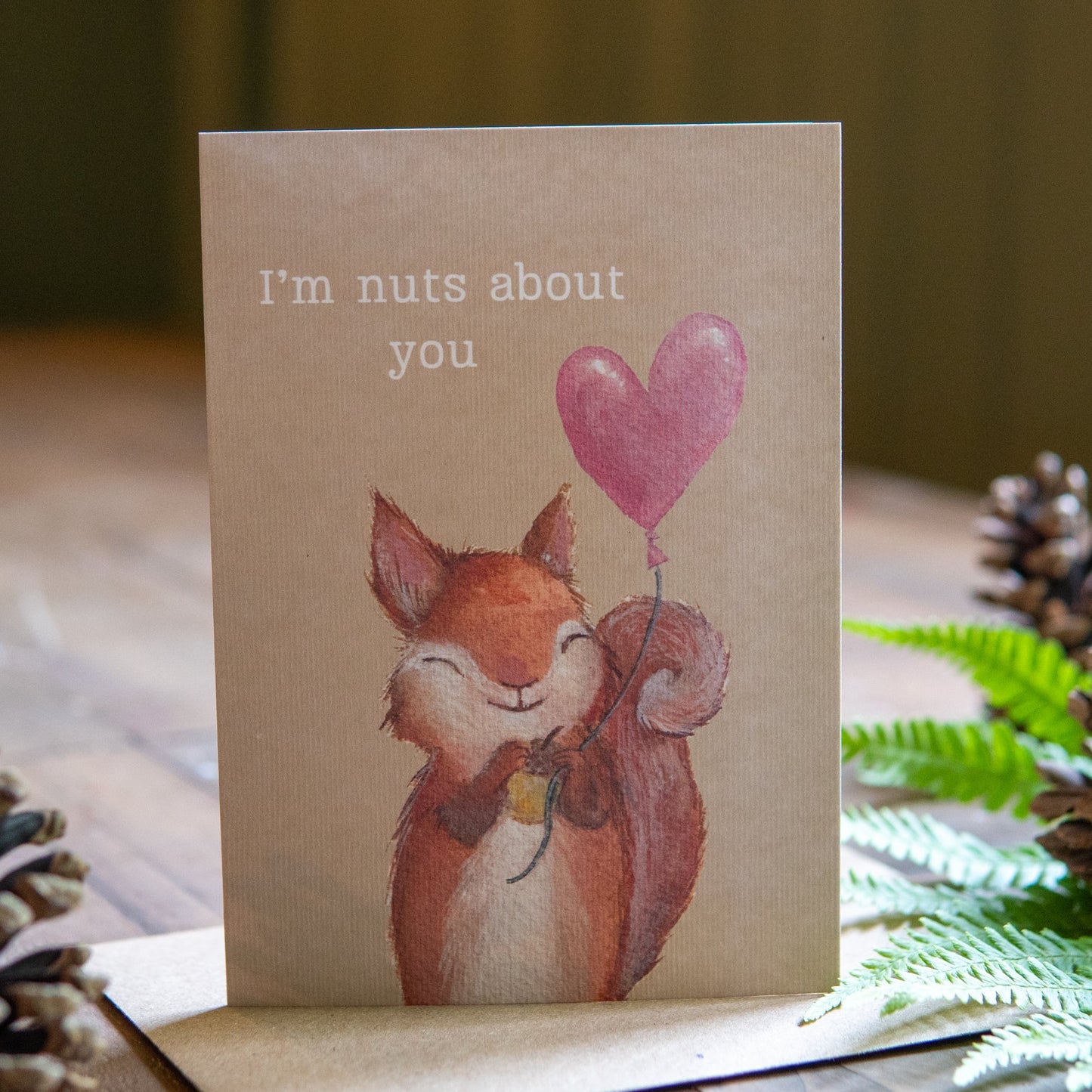 Card with a fox holding a heart-shaped balloon and text 'I'm nuts about you' on a wooden surface with decorative elements.