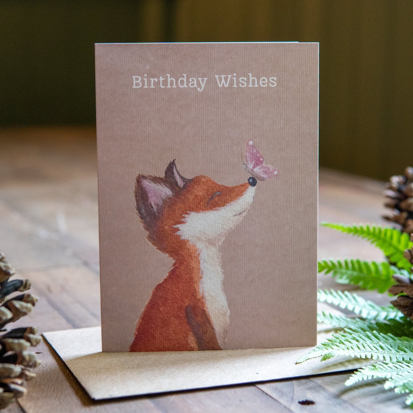 Greeting card with a fox illustration and 'Birthday Wishes' text, surrounded by pine cones and ferns on a wooden surface.