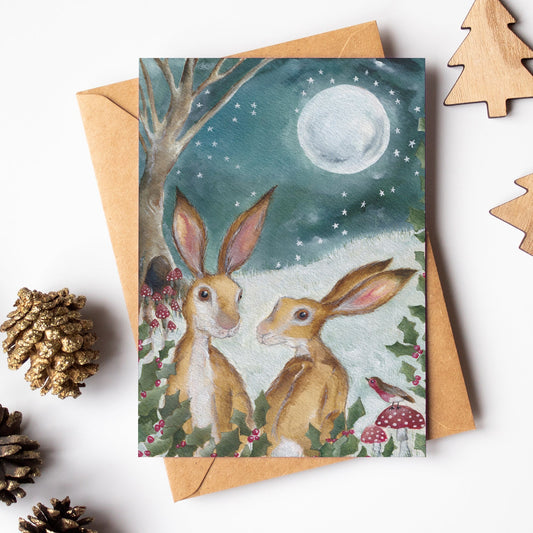 Greeting card with two rabbits under a moonlit sky, surrounded by pinecones and wooden trees on a white background.