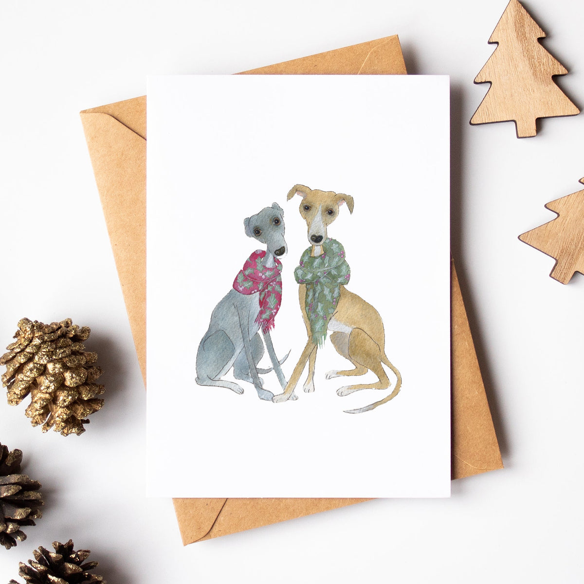 A white greeting card featuring a watercolor illustration of two greyhounds wearing Christmas sweaters, displayed on a kraft paper envelope with pinecones and wooden tree cutouts on a white background.
