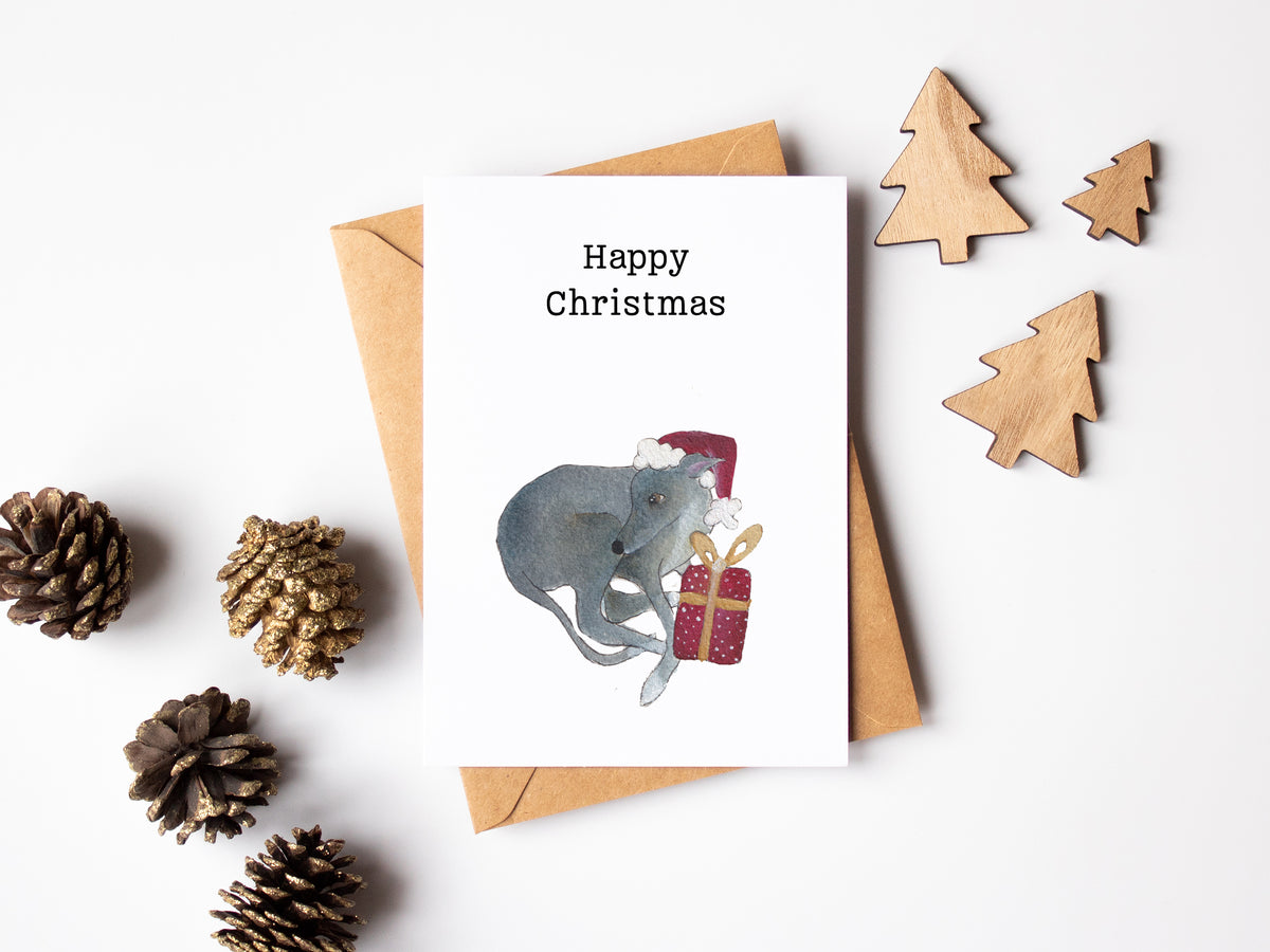 A Christmas greeting card with a watercolor illustration of a gray cat wearing a red Santa hat and hugging a red gift box, placed on top of a brown kraft envelope. The card is photographed in a styled flat lay on a white background with wooden tree cutouts, pinecones, and a small wrapped gift.